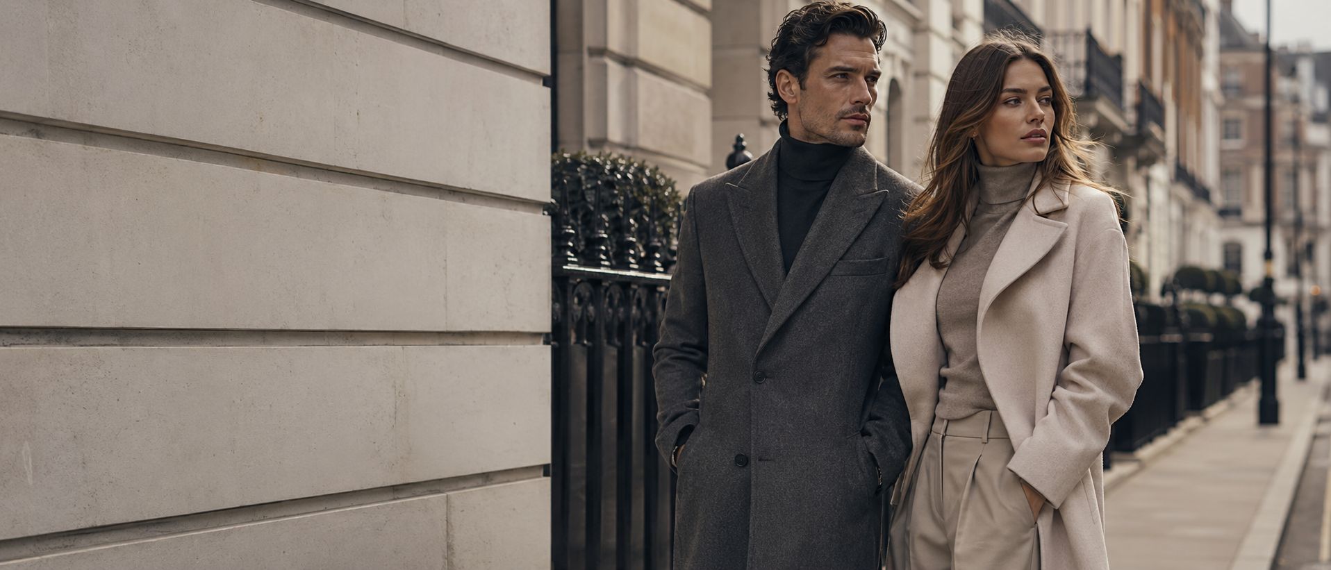 A man in a grey overcoat and a woman in a cream coat walking together along a city street